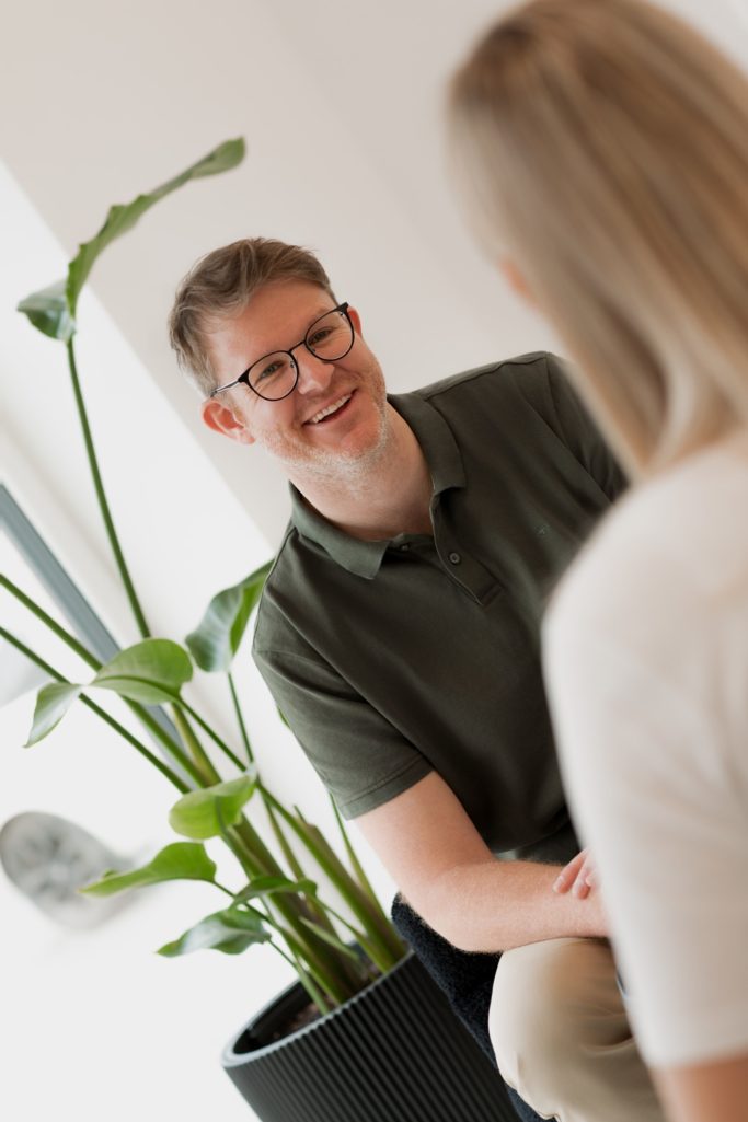 Mann mit Brille sitzt lächelnd neben einer Pflanze, Blickrichtung auf eine Person.